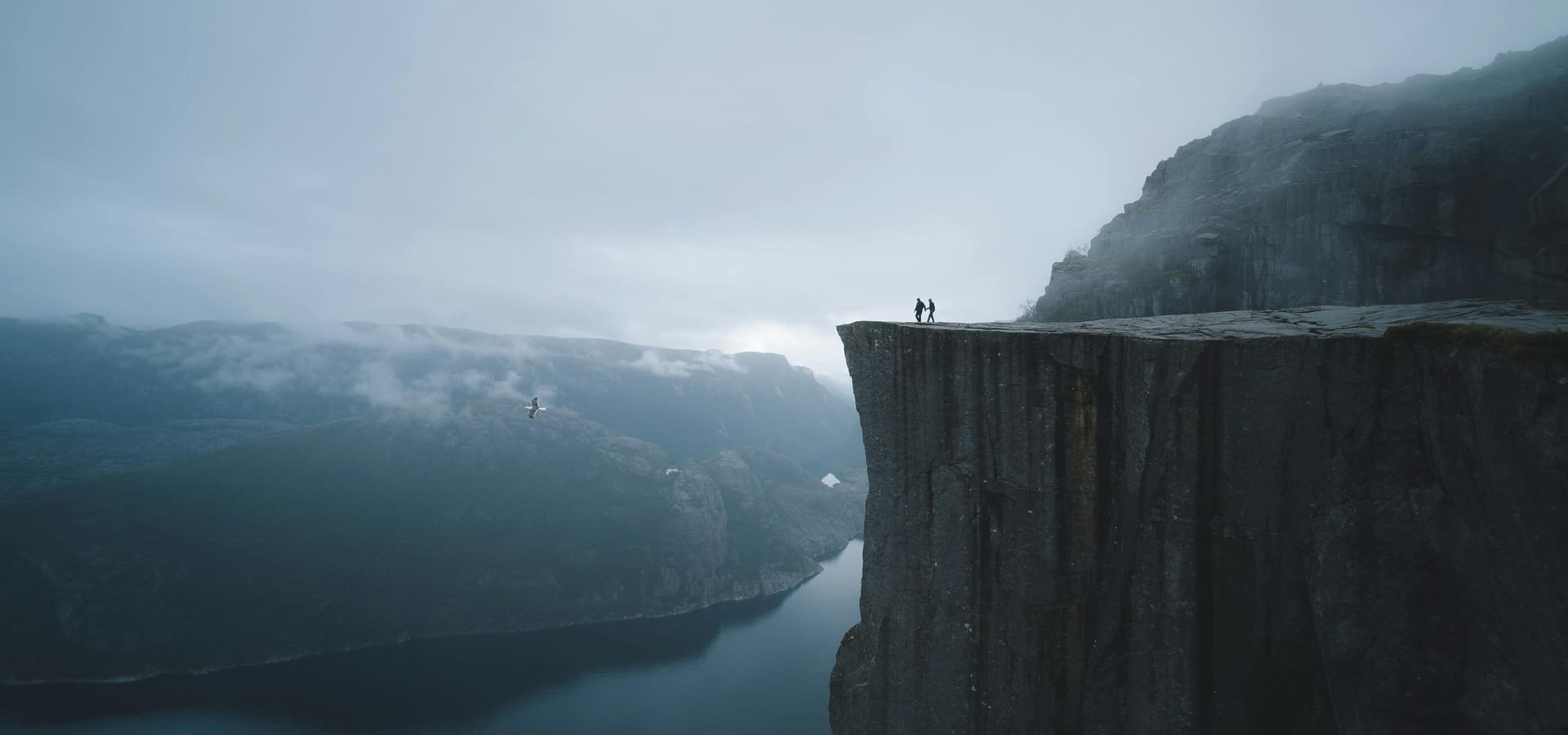 Two travellers standing on a dramatic cliff edge overlooking a fjord